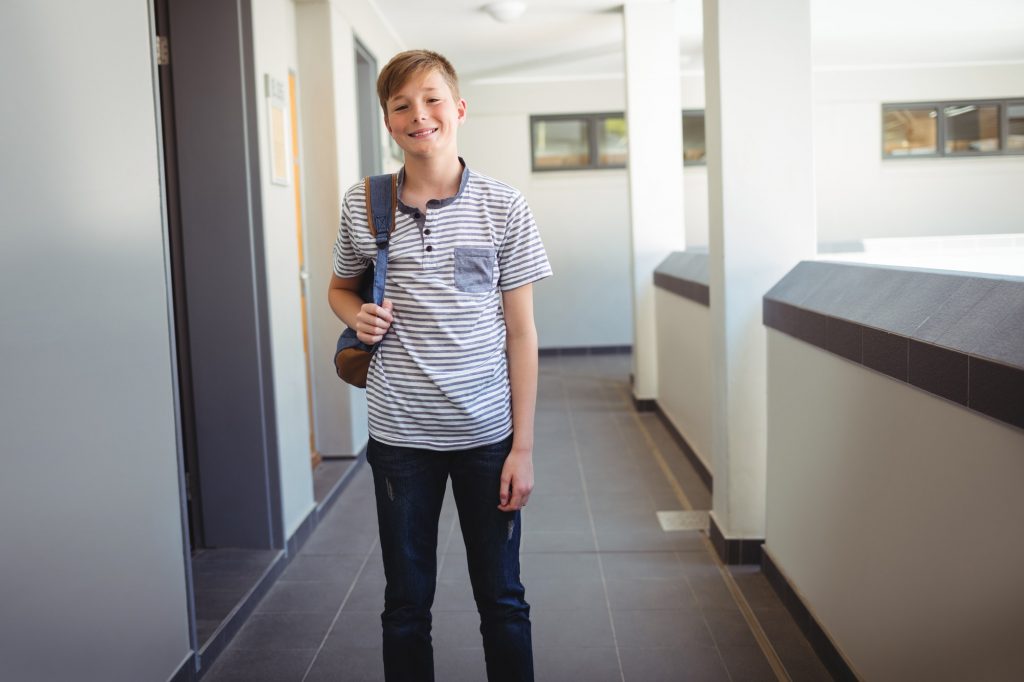 Smiling schoolboy standing with schoolbag in corridor at school